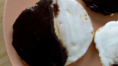 A close-up of a black and white cookie on an orange plate, perfect for anyone searching for a classic Black and White Cookies recipe. The cookie is half-covered in chocolate icing and half in white, with another partially visible nearby.