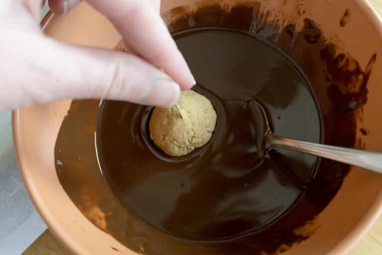 A hand dips a peanut butter ball held with a toothpick into a bowl of melted chocolate, with a spoon resting in the chocolate—perfect for making this Easy Buckeyes Recipe.