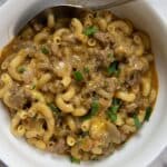 A white bowl filled with Homemade Hamburger Helper—macaroni pasta mixed with ground beef, melted cheese, and garnished with chopped parsley. A spoon rests inside the bowl.