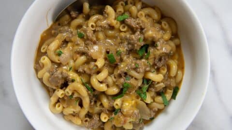 A white bowl filled with Homemade Hamburger Helper—macaroni pasta mixed with ground beef, melted cheese, and garnished with chopped parsley. A spoon rests inside the bowl.