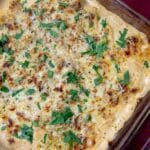 A baked casserole of Spicy Italian Sausage Dip in a glass dish, topped with melted cheese and chopped fresh parsley, on a red tablecloth.