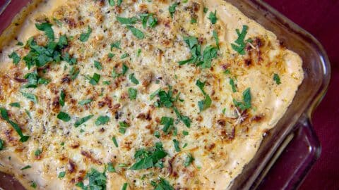 A baked casserole of Spicy Italian Sausage Dip in a glass dish, topped with melted cheese and chopped fresh parsley, on a red tablecloth.
