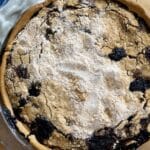 A close-up of a Shoofly Pie with a golden-brown, slightly cracked crust and patches of dark fruit filling, dusted with powdered sugar, in a clear pie dish next to a blue-striped kitchen towel.