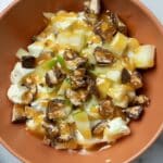 A bowl of Apple Snickers Salad Recipe features chopped green apples, diced chocolate candy bars, and a drizzle of caramel over a creamy white base. A metal spoon rests beside the bowl on a marble surface.