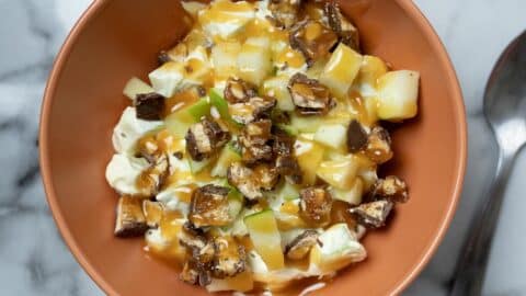 A bowl of Apple Snickers Salad Recipe features chopped green apples, diced chocolate candy bars, and a drizzle of caramel over a creamy white base. A metal spoon rests beside the bowl on a marble surface.