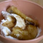 A peach-colored bowl with vanilla ice cream topped with caramelized bananas and brown syrup on a wooden surface, inspired by a classic Bananas Foster recipe.