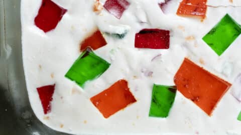 Close-up of a dessert featuring red, green, and orange gelatin cubes suspended in a creamy white mixture, likely set in a glass dish—perfect for showcasing a classic Broken Glass Cake Recipe.