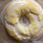 A round pastry with a hole in the center, covered in glossy white glaze, sits on brown parchment paper. One section is bitten off, revealing a flaky texture inside—just like a classic Danish Kringle recipe brought to life.