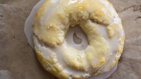 A round pastry with a hole in the center, covered in glossy white glaze, sits on brown parchment paper. One section is bitten off, revealing a flaky texture inside—just like a classic Danish Kringle recipe brought to life.