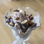 A glass dish filled with chocolate ice cream, creamy whipped cream, chopped nuts, and rich chocolate sauce sits on a wooden surface—just like a classic Tin Roof Sundae Recipe brought to life.