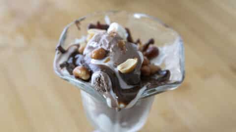 A glass dish filled with chocolate ice cream, creamy whipped cream, chopped nuts, and rich chocolate sauce sits on a wooden surface—just like a classic Tin Roof Sundae Recipe brought to life.