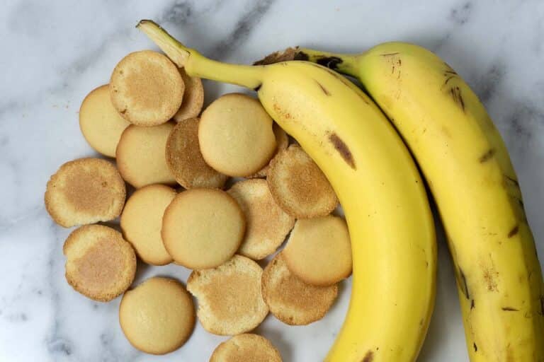 Two ripe yellow bananas laying next to a pile of round, golden-brown vanilla wafer cookies on a white marble surface.