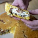 A person holds a slice of savory Jalapeño Popper Cornbread, stuffed with cheese, herbs, and mushrooms, while the rest of the cornbread sits on a plate in the background on a wooden surface.