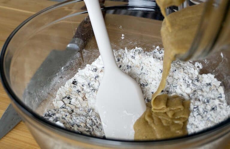 A white spatula sits in a glass bowl with flour and chocolate chips as thick brown batter from The Best Homemade Cowboy Cookies Recipe is poured in from the side.