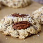 A close-up of an oatmeal cookie topped with a pecan half, resting on parchment paper. Shredded coconut is sprinkled on the cookie, echoing The Best Homemade Cowboy Cookies Recipe, with more cookies blurred in the background.