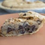 A close-up of a slice of chocolate chip cookie pie inspired by a classic Kentucky Derby Pie recipe on a peach-colored plate, showing a thick, gooey filling with visible chocolate chips and a golden, crumbly crust. The rest of the pie is blurred in the background.
