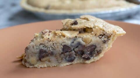 A close-up of a slice of chocolate chip cookie pie inspired by a classic Kentucky Derby Pie recipe on a peach-colored plate, showing a thick, gooey filling with visible chocolate chips and a golden, crumbly crust. The rest of the pie is blurred in the background.