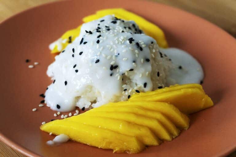 A serving of mango sticky rice on a brown plate, topped with coconut sauce and sprinkled with black and white sesame seeds, with fresh sliced mango on the side.