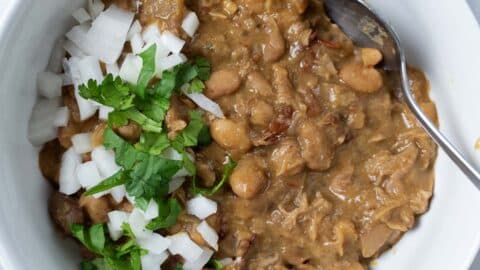 A bowl of Crock Pot Pinto Beans, refried and topped with chopped white onions and fresh cilantro, with a spoon resting inside the bowl.
