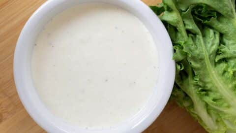A white bowl filled with creamy buttermilk ranch dressing, placed on a wooden surface next to fresh green lettuce leaves.