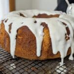 A freshly baked bundt cake made from a Sundrop Cake Recipe with thick white icing drizzling down the sides sits on a wire rack, as a spoon adds more icing to the top.