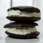 Two chocolate whoopie pies with a thick layer of white cream filling are stacked on a white marble surface, showcasing the irresistible results of this classic whoopie pie recipe.