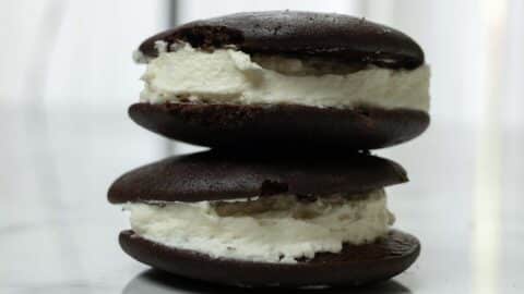 Two chocolate whoopie pies with a thick layer of white cream filling are stacked on a white marble surface, showcasing the irresistible results of this classic whoopie pie recipe.
