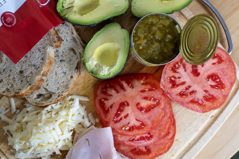 A wooden cutting board holds all the fixings for an Albuquerque Turkey Sandwich: sliced bread, halved avocados, tomato slices, shredded cheese, deli ham, and an open can of diced green chilies.