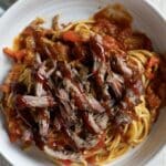 A white bowl filled with BBQ Spaghetti—tender spaghetti topped with shredded barbecue beef and chunky tomato sauce—sits on a light-colored tablecloth, with a fork visible to the side.