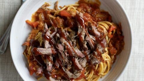 A white bowl filled with BBQ Spaghetti—tender spaghetti topped with shredded barbecue beef and chunky tomato sauce—sits on a light-colored tablecloth, with a fork visible to the side.