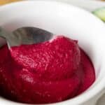 A close-up of a spoon scooping vibrant pink beet hummus from a white bowl, showcasing this delicious beet hummus recipe, with a slice of cucumber visible in the background.
