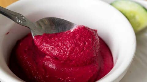 A close-up of a spoon scooping vibrant pink beet hummus from a white bowl, showcasing this delicious beet hummus recipe, with a slice of cucumber visible in the background.