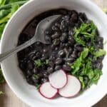 A white bowl filled with cooked black beans, garnished with chopped cilantro and three radish slices, with a spoon resting in the bowl. Fresh cilantro sits beside the bowl, completing this vibrant Black Beans Recipe presentation.