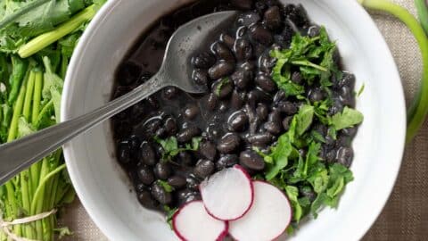 A white bowl filled with cooked black beans, garnished with chopped cilantro and three radish slices, with a spoon resting in the bowl. Fresh cilantro sits beside the bowl, completing this vibrant Black Beans Recipe presentation.
