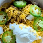 A bowl of chili inspired by a classic Frito Pie recipe, topped with shredded cheddar cheese, sour cream, sliced jalapeños, and chopped white onions. The vibrant ingredients are arranged on top in a white bowl.