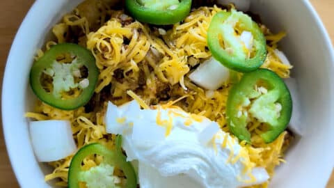 A bowl of chili inspired by a classic Frito Pie recipe, topped with shredded cheddar cheese, sour cream, sliced jalapeños, and chopped white onions. The vibrant ingredients are arranged on top in a white bowl.