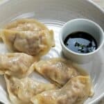 Five steamed dumplings from a classic Mandu recipe are arranged on a white plate next to a small cup of soy sauce with chopped green onions. Chopsticks rest on the edge, set against a beige woven fabric background.