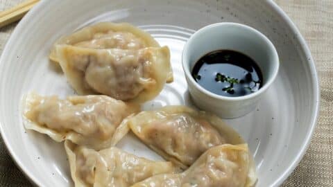 Five steamed dumplings from a classic Mandu recipe are arranged on a white plate next to a small cup of soy sauce with chopped green onions. Chopsticks rest on the edge, set against a beige woven fabric background.