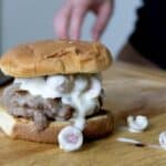 An Olive Burger featuring a juicy beef patty and grapes covered in white sauce is served on a wooden cutting board. A hand rests behind the burger, with extra grapes scattered nearby for a unique twist.
