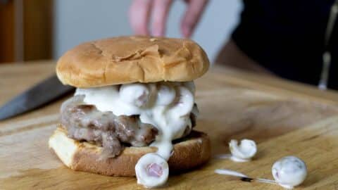 An Olive Burger featuring a juicy beef patty and grapes covered in white sauce is served on a wooden cutting board. A hand rests behind the burger, with extra grapes scattered nearby for a unique twist.