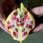 Two hands hold a yellow pastry or dessert cut in half, revealing a colorful filling with strawberry, kiwi, and white cream inside—resembling the vibrant layers in a Mango Lassi Yogurt Cluster Recipe. The background is blurred.