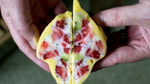 Two hands hold a yellow pastry or dessert cut in half, revealing a colorful filling with strawberry, kiwi, and white cream inside—resembling the vibrant layers in a Mango Lassi Yogurt Cluster Recipe. The background is blurred.