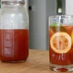 A glass of iced tea with a lemon slice and mint leaf sits on a wooden table next to a mason jar filled with more iced tea, hinting at a refreshing How To Make Cold Brew Iced Strawberry Tea Recipe. A blurred background shows part of a plant and a doorway.