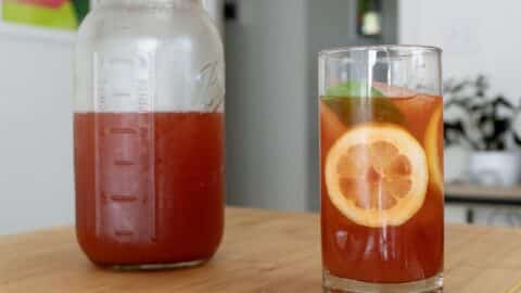 A glass of iced tea with a lemon slice and mint leaf sits on a wooden table next to a mason jar filled with more iced tea, hinting at a refreshing How To Make Cold Brew Iced Strawberry Tea Recipe. A blurred background shows part of a plant and a doorway.