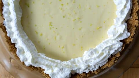 A classic Key lime pie in a glass dish features a graham cracker crust, creamy yellow filling, whipped cream piped around the edge, and a sprinkle of fresh lime zest on top.