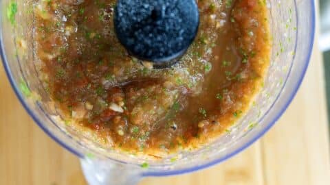 A close-up, top-down view of a food processor containing freshly blended salsa with visible pieces of tomatoes, onions, garlic, and cilantro—perfect for making a delicious Salsa Tatemada Recipe. The processor rests on a wooden surface.