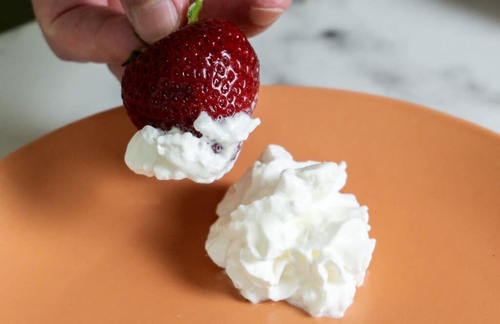 A strawberry being dunked into whipped cream on a plate.