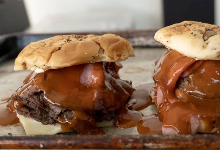 Two North Shore Beef sandwiches with thick slices of meat, melted cheese, onions, and barbecue sauce overflowing from sesame seed buns are served on a baking sheet.