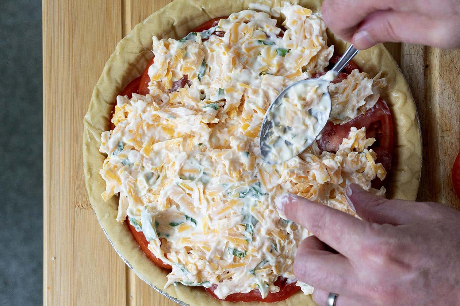 A person spreads a creamy cheese mixture over sliced tomatoes in an unbaked pie crust using a spoon, following a classic Tomato Pie Recipe on a wooden surface.