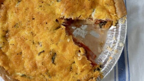 A baked savory Tomato Pie Recipe features a golden, cheesy crust in a foil pan, with a slice removed to reveal its creamy tomato filling. The pie rests on a white cloth with blue stripes.